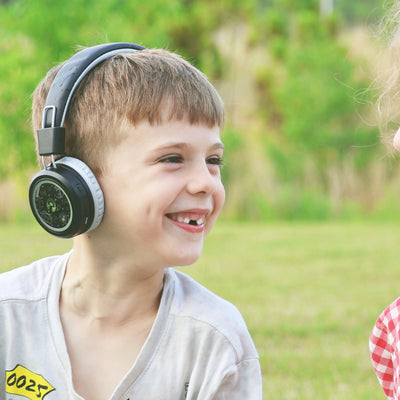 Child wearing MEE audio KidJamz Share Bluetooth Wireless Headphones for Kids with Multipoint, LED Lights, 85dB Volume Limiter, Microphone, 40h battery; Childrens Headset for School/iPhone/iPad/Tablet  in a park setting