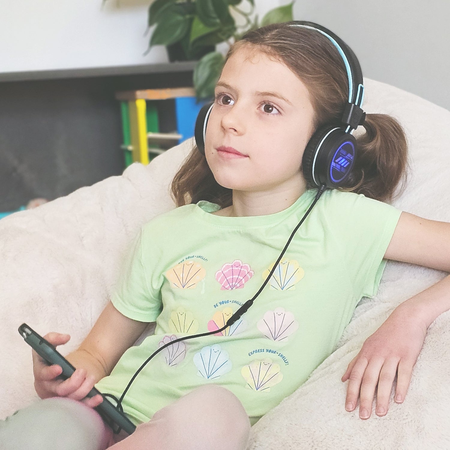A young girl lounging on a couch while wearing large headphones connected to a smartphone, looking thoughtful. there's a potted plant in the background.