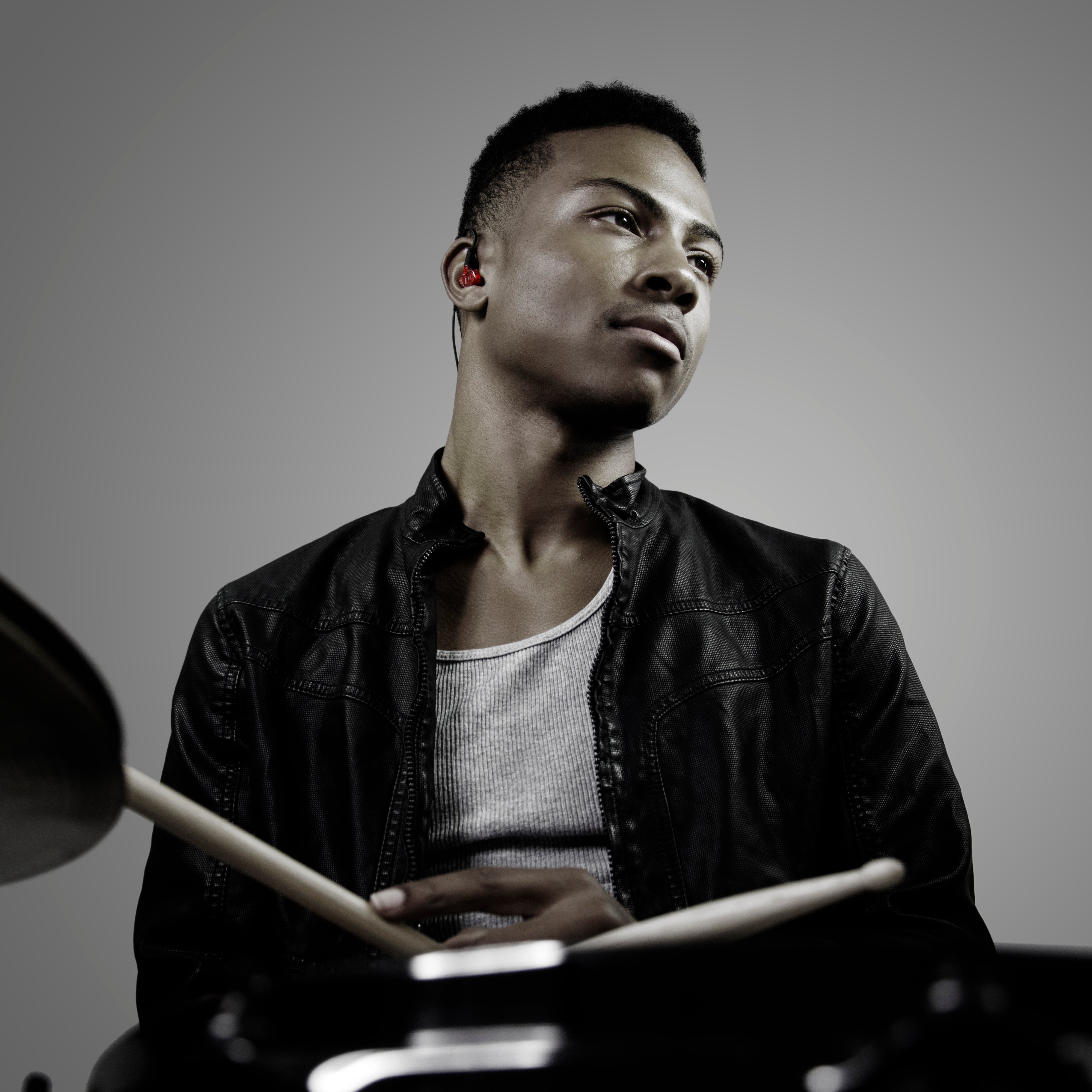 A young male drummer wearing a leather jacket sits behind a drum kit, looking to his left with a focused expression. he holds drumsticks and wears earphones.