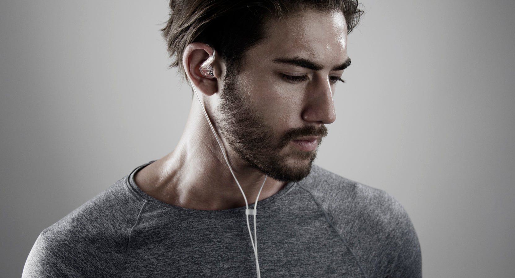 A young man with stubble wearing a gray t-shirt and white earphones, looking downwards thoughtfully against a neutral gray background.