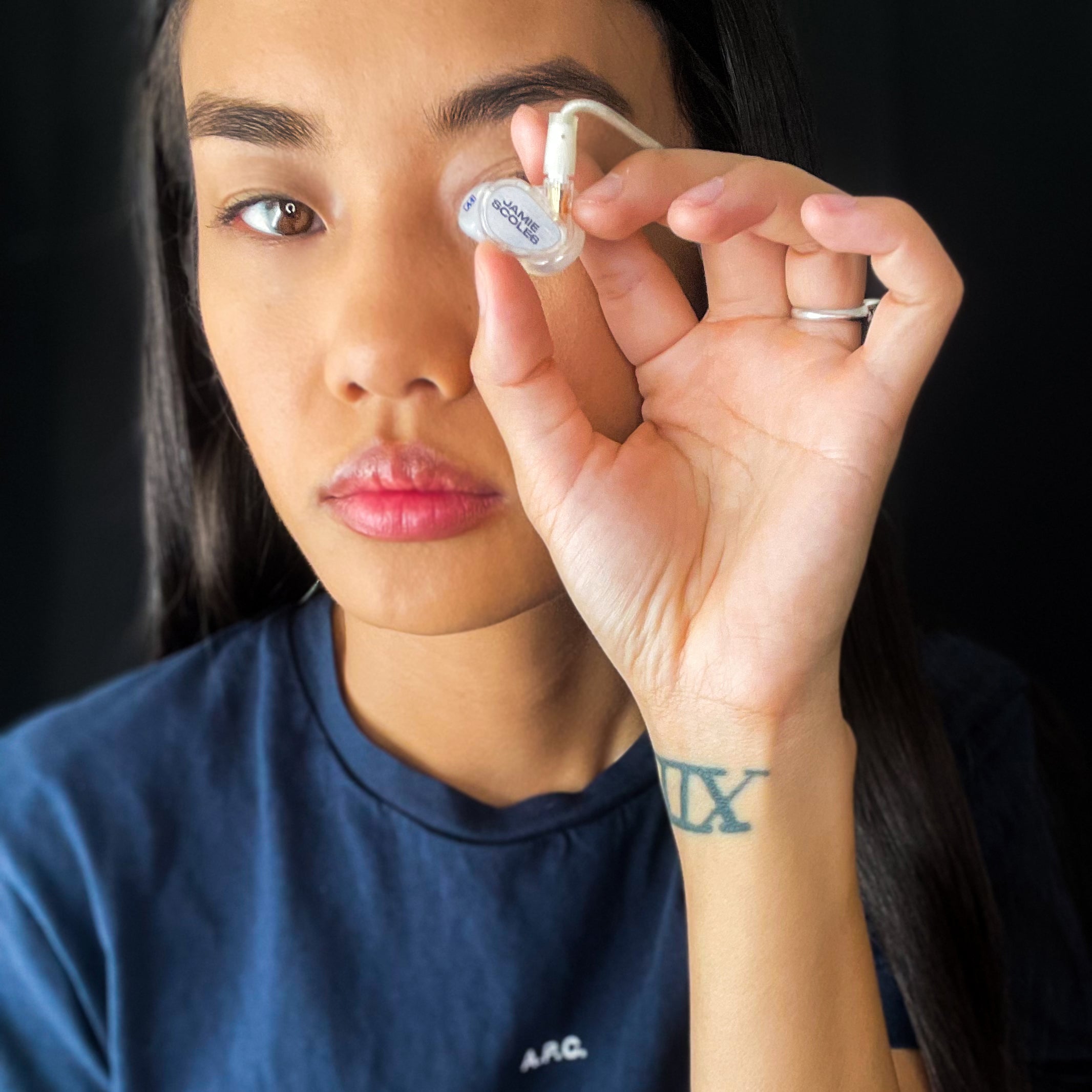 A woman holding a small white earbud close to her eye for inspection, with a focused expression, wearing a dark blue t-shirt with a visible tattoo on her forearm.