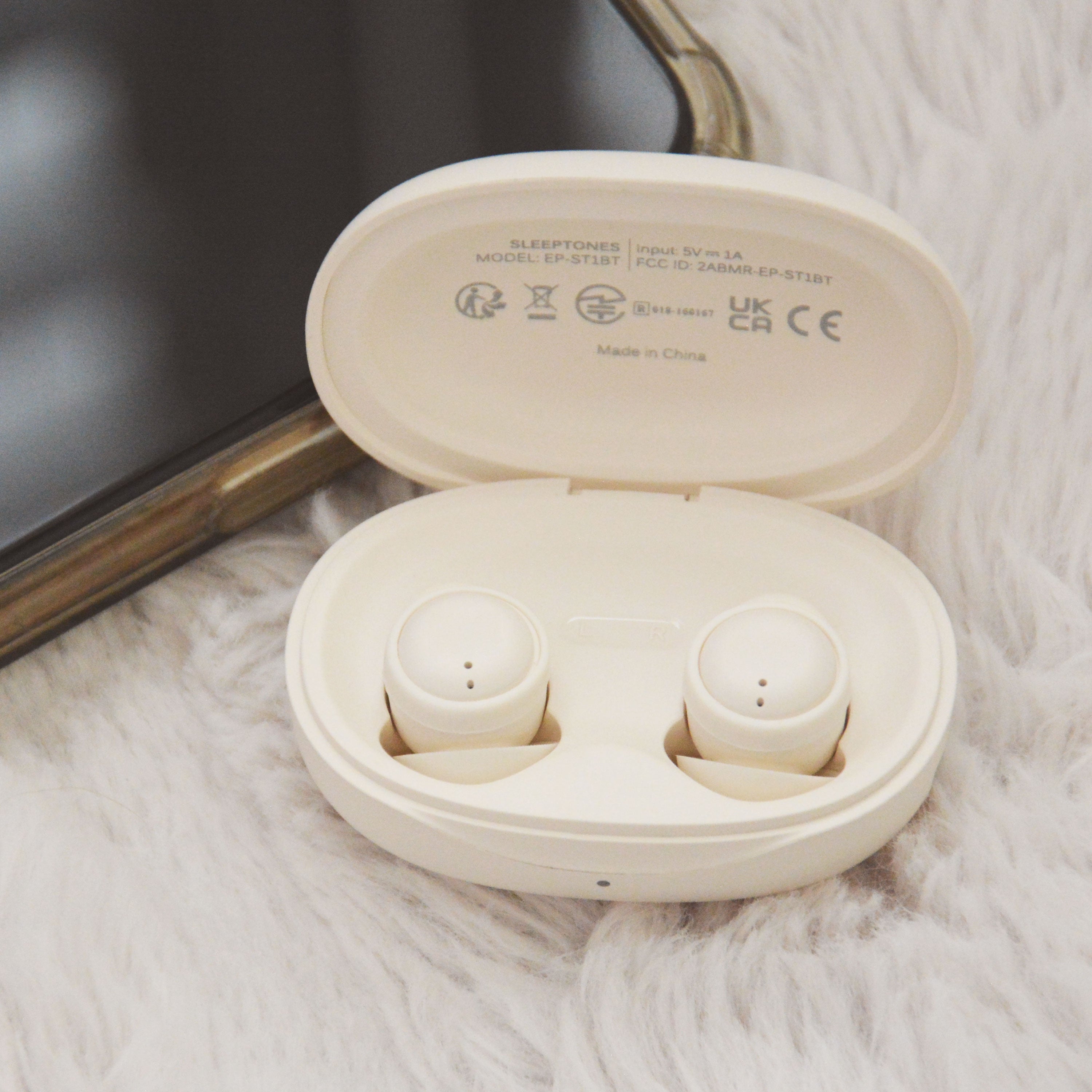 Open cream-colored wireless SLEEPTONES earbuds charging case with two earbuds inside, photographed on a white faux-fur surface next to a dark rectangular object.
