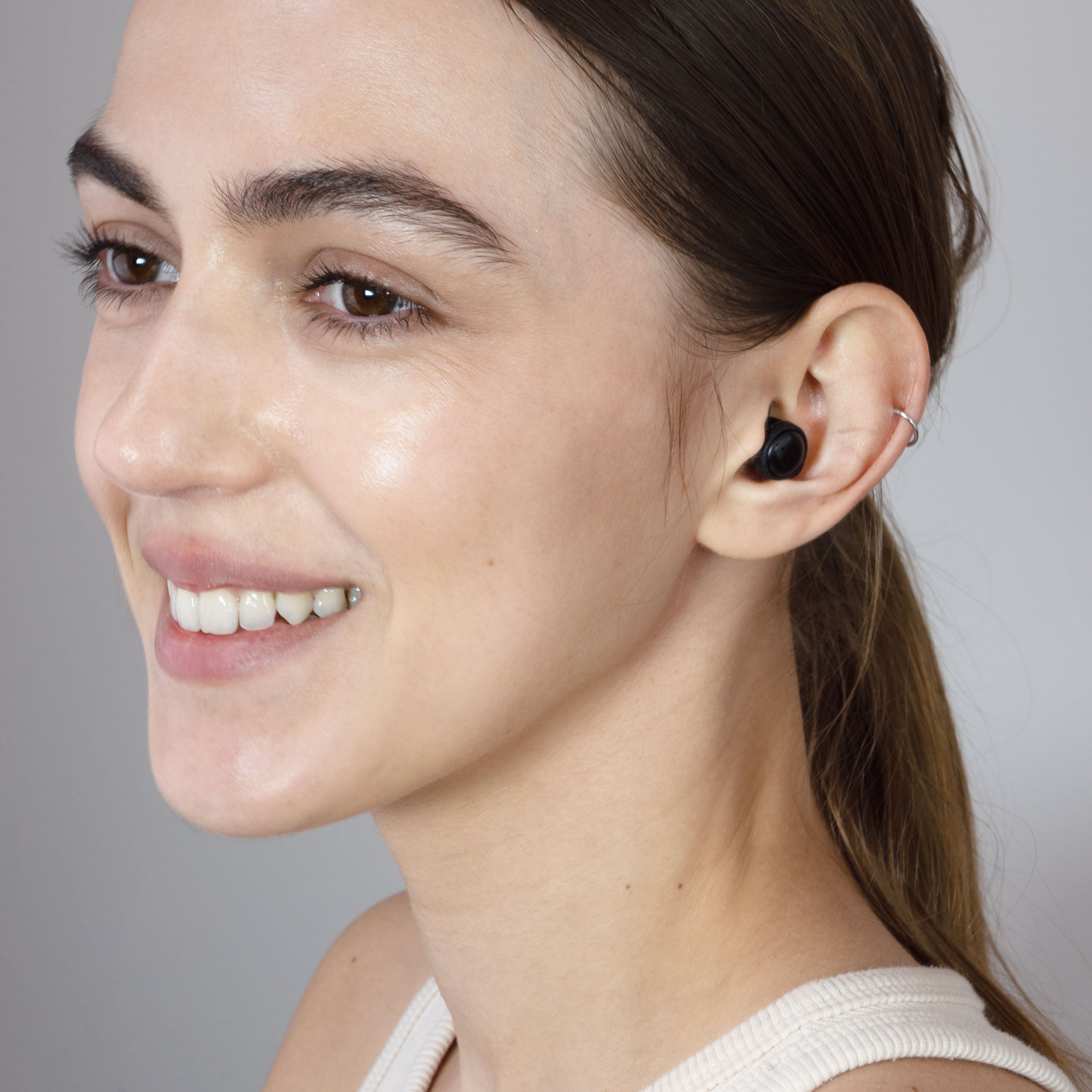 A close-up side profile of a smiling woman with her hair tied back, wearing a small black in-ear wireless SLEEPTONES earbud in her ear against a plain gray background.