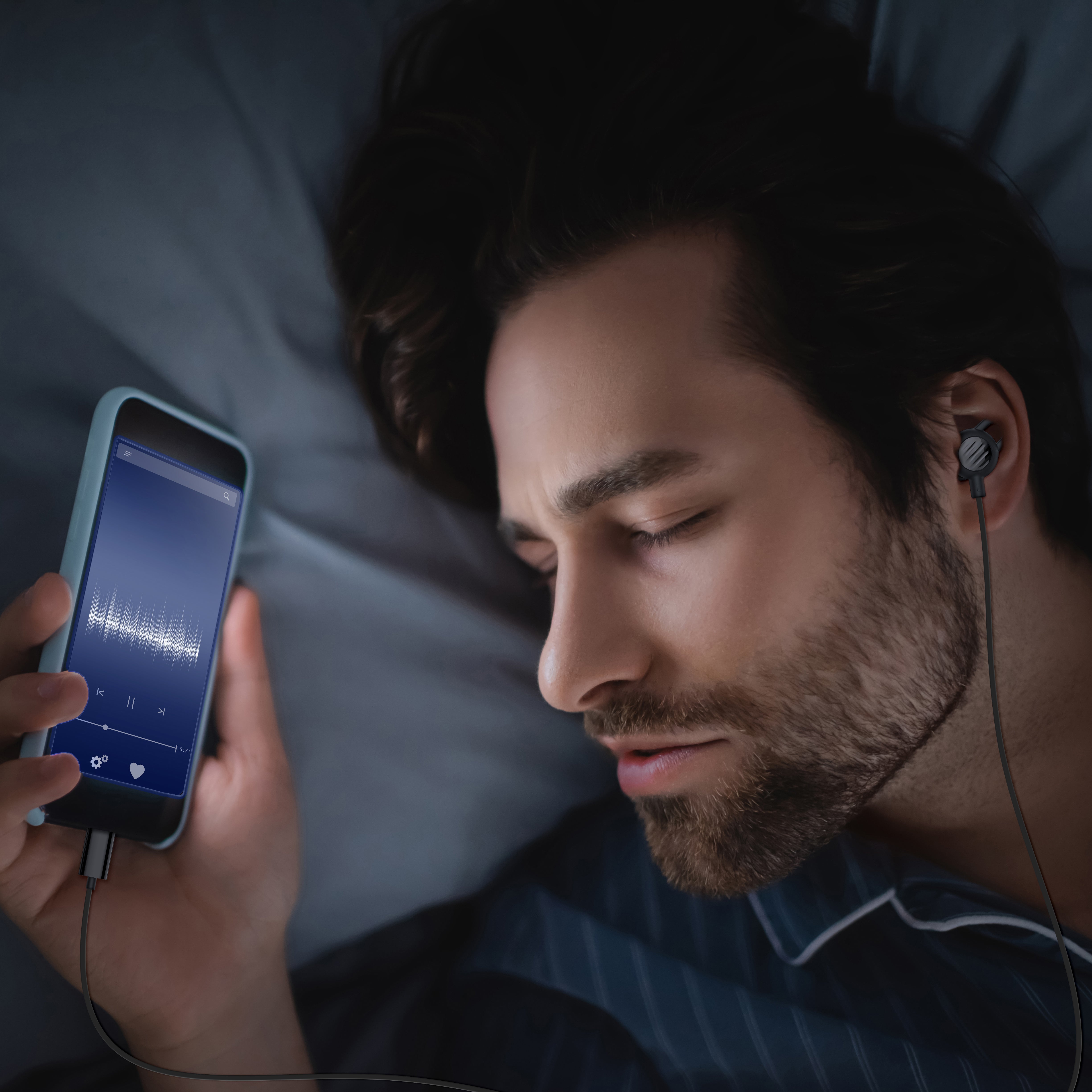Man sleeping using SleepTones Wired Silicone Sleeping Earbuds while lying in bed using a smartphone listening to white noise in a dimly lit room.
