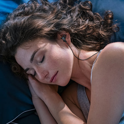 Woman lying down with eyes closed, wearing MEE audio SleepTones Wired Sleeping Earbuds reduces pressure points so you can sleep soundly.