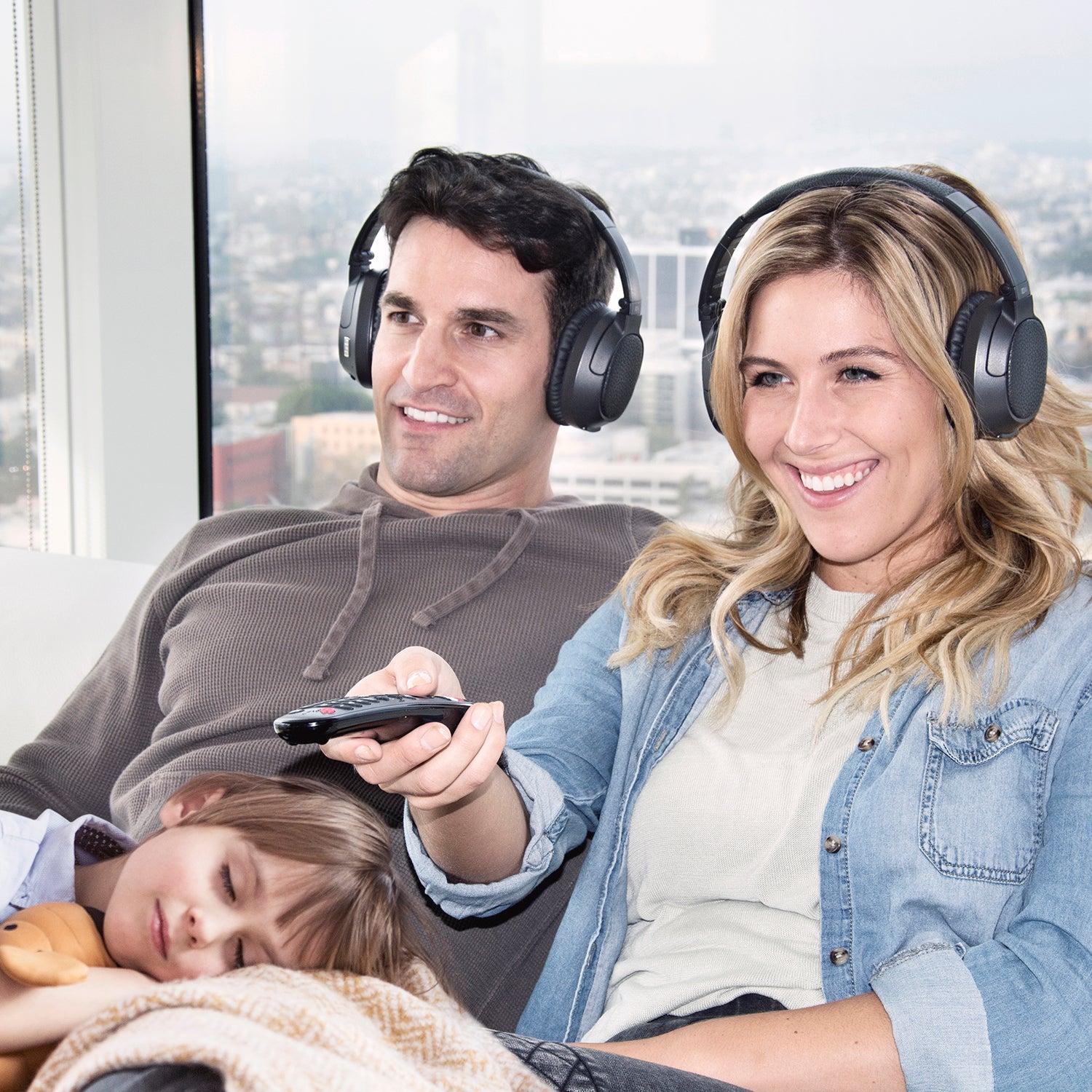 A happy family enjoying time together in a sunlit room; a man and a woman, both wearing headphones, smile warmly while a child sleeps in the woman's lap, holding a remote control.