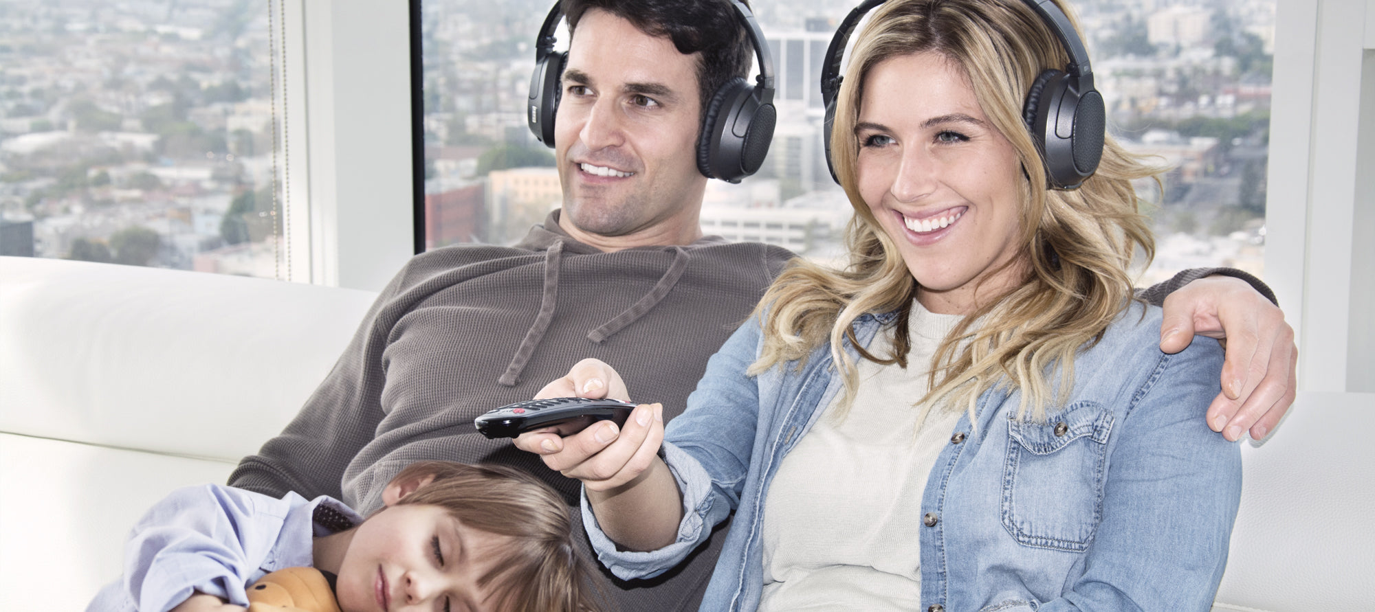 A cheerful young woman and a man wearing headphones sit on a couch, smiling at the camera, while a young child sleeps on the woman's lap. the man holds a remote control. bright cityscape visible through the window behind them.