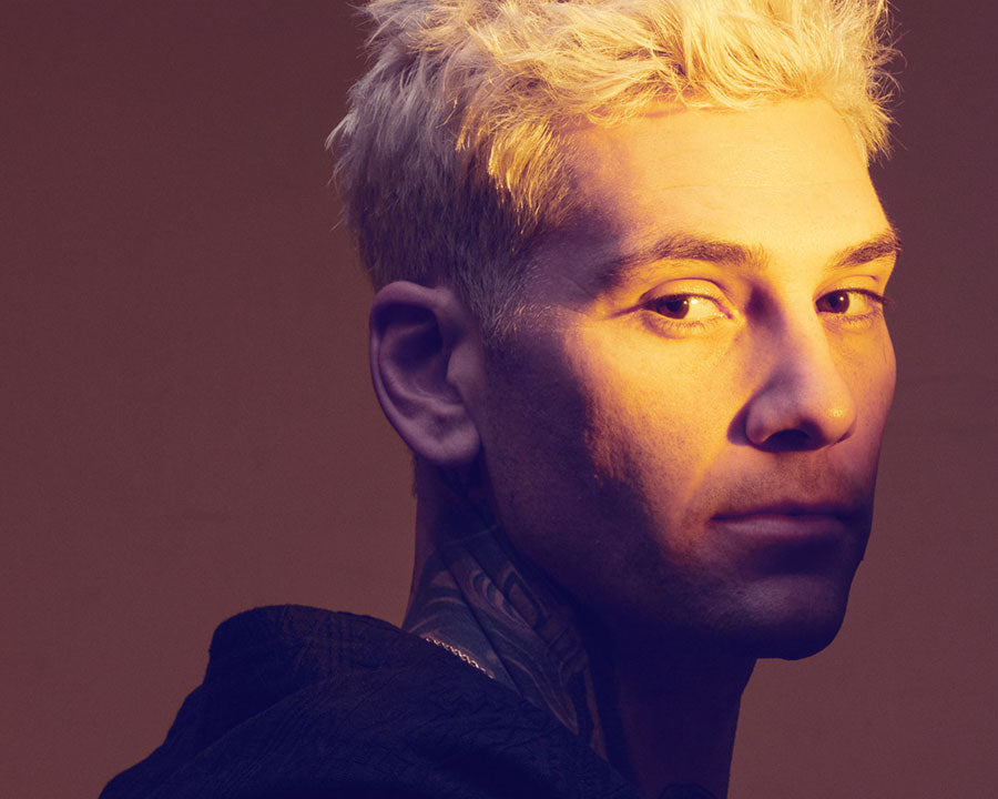 Side profile of a man with platinum blonde hair and visible tattoos on his neck, illuminated by warm golden light against a plain background.