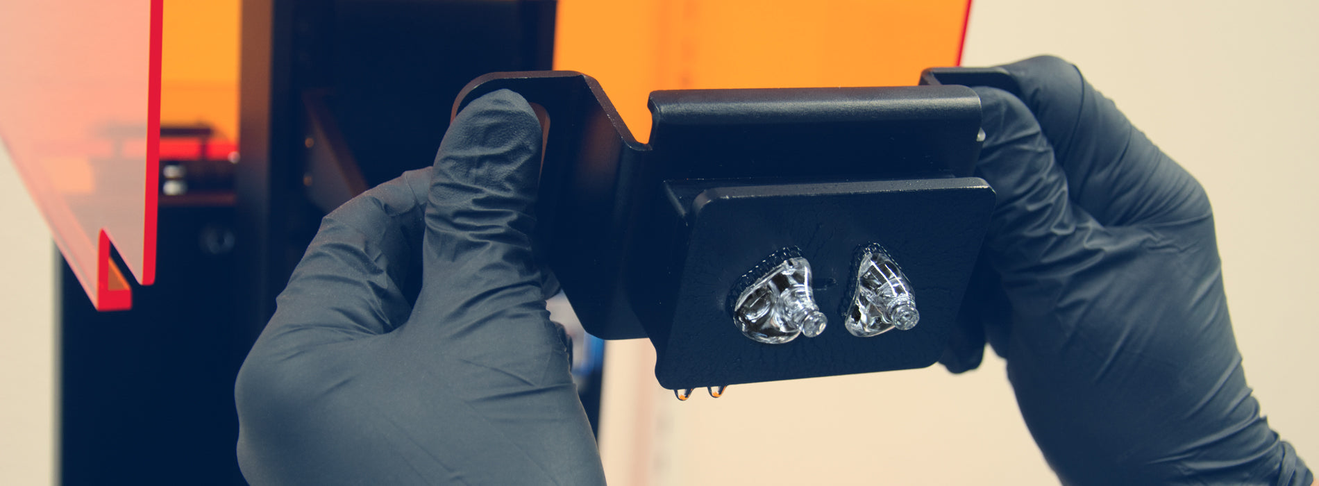 Hands in black gloves holding a gemstone under a microscope for examination against an orange-lit background.