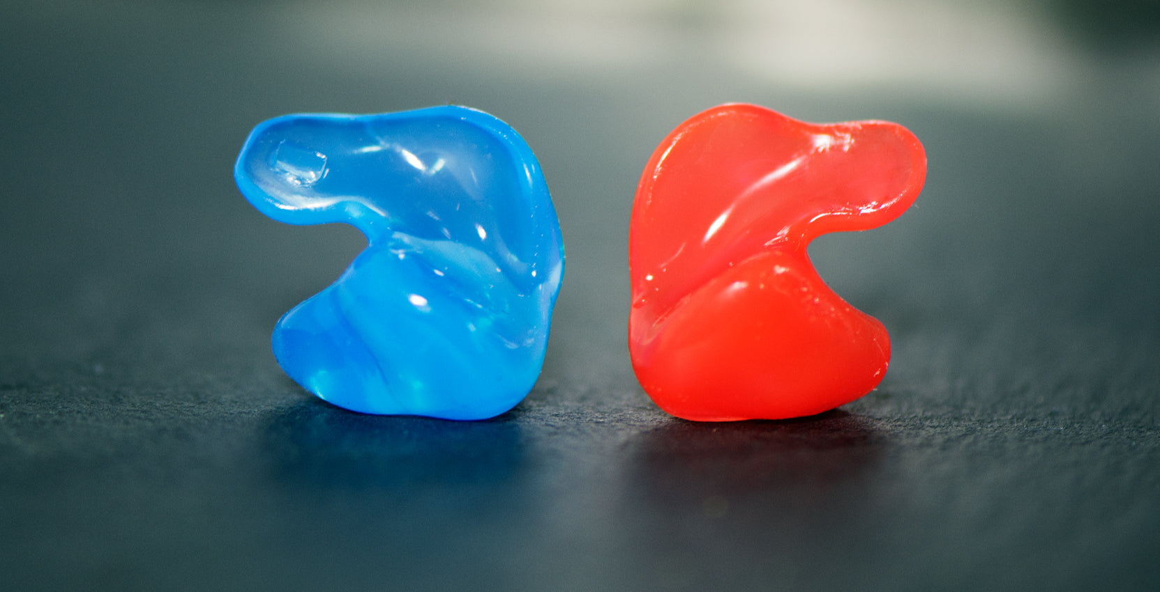 Two vibrant, translucent earplugs, one blue and one red, placed on a gray surface, captured in close-up showcasing their squishy texture.