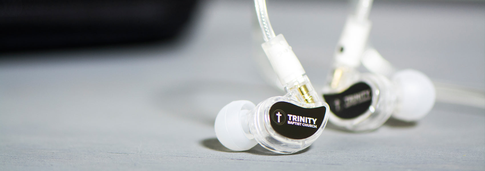 Close-up of clear, in-ear headphones with white cables and logo "trinity audio engineering" on a gray surface.
