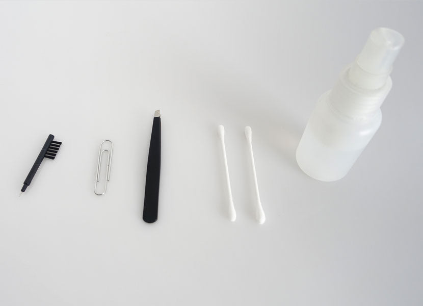 An array of office supplies on a white background, including a pair of black plastic tweezers, a metal paperclip, a black pen, two cotton swabs, and a small, white, spray bottle.