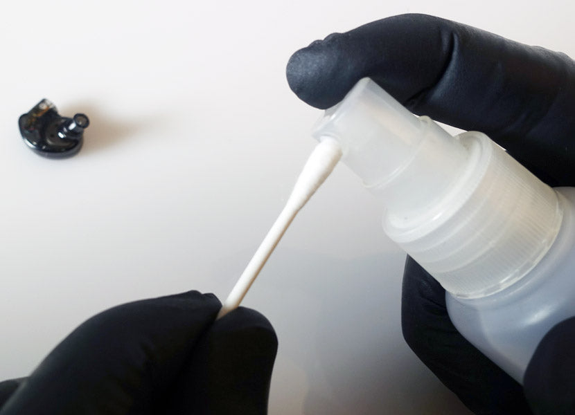 Gloved hands cleaning an earbud with a cotton swab dipped in cleaner from a squeeze bottle, with the detached earbud piece lying nearby.