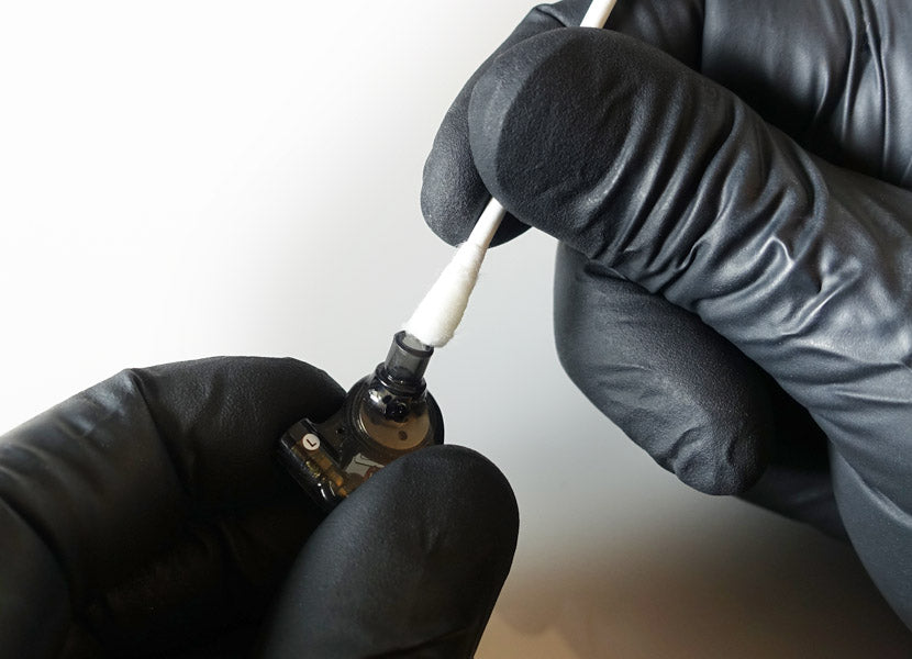 Hands in black gloves applying a cleaning solution with a cotton swab to a small electronic device component.