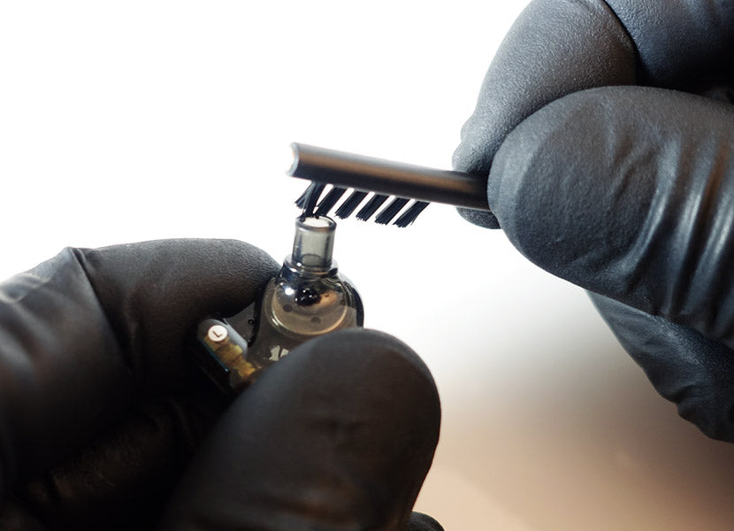 Hands wearing black gloves cleaning a small electronic device with a brush, focused on precision and detail.