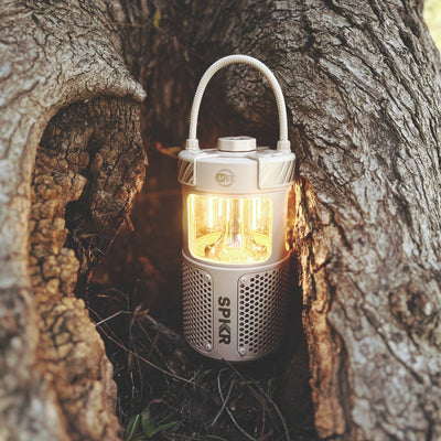 Beige lightSPKR Bluetooth Wireless Speaker with Camping Lantern tucked near a tree