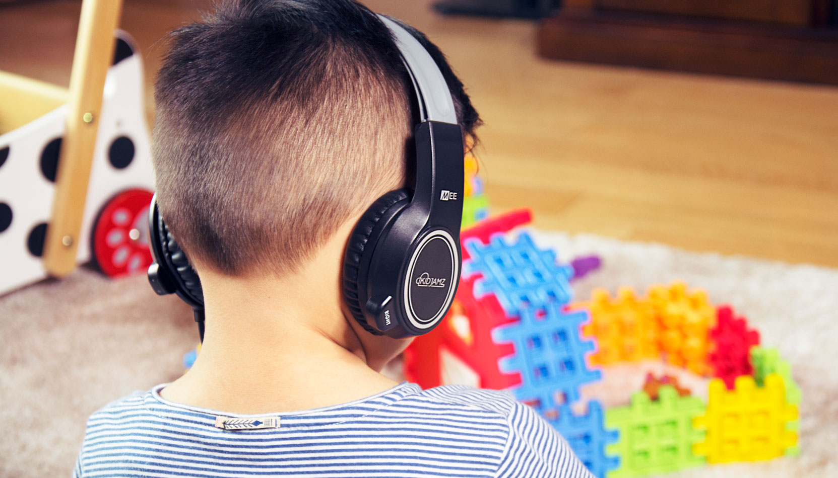 Young boy wearing headphones while playing with colorful blocks on a carpeted floor, with toys scattered around.