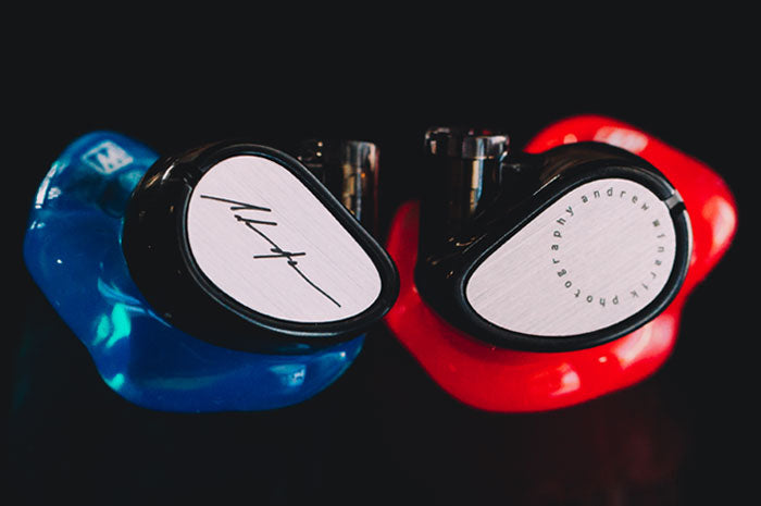 Two custom in-ear monitors, one with a blue shell and another with a red shell, each featuring a metallic faceplate with engraved designs, on a black background.