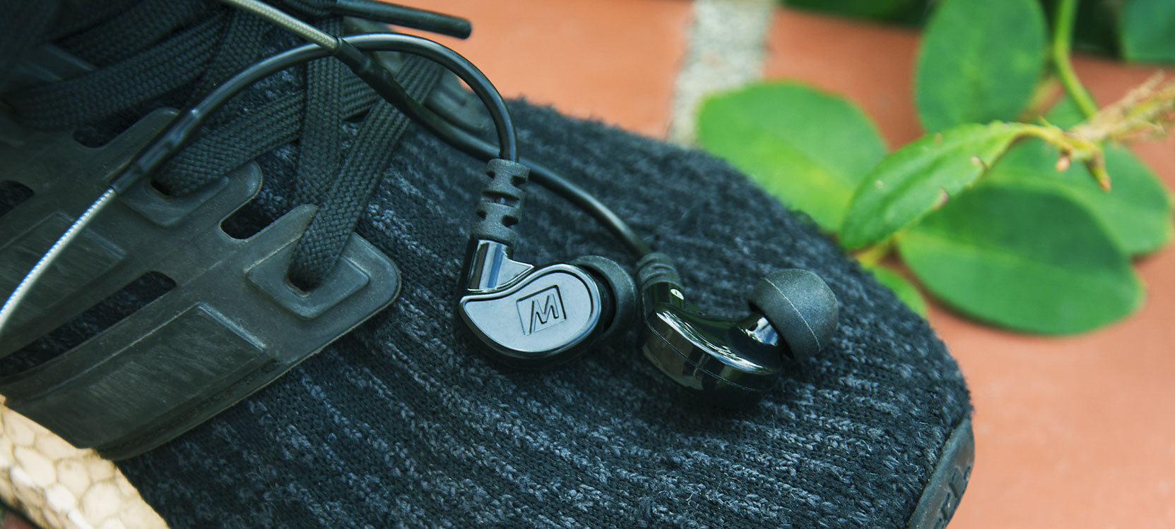 A pair of in-ear headphones with a logo rests on the brim of a woven hat, next to a sandal and a green plant, all on a coral-colored surface.