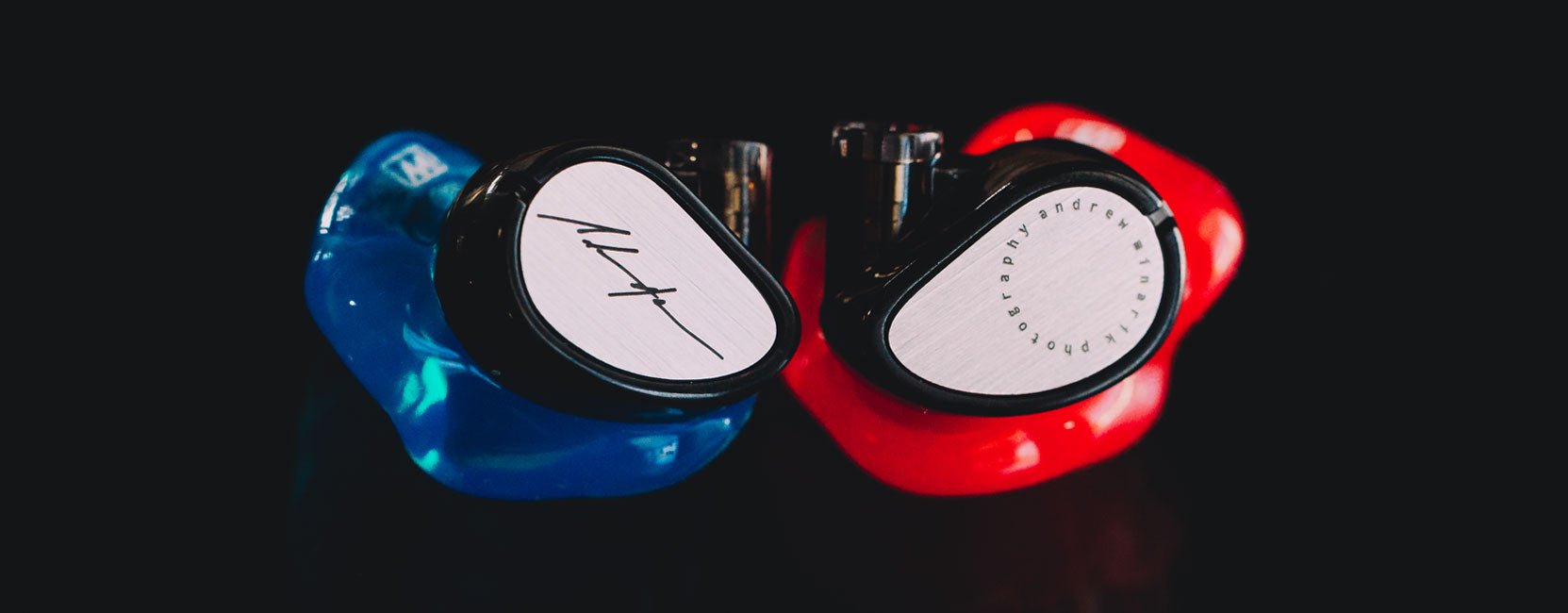Two pairs of custom in-ear monitors, one in blue and one in red, both featuring distinctive logo designs on a reflective black surface.
