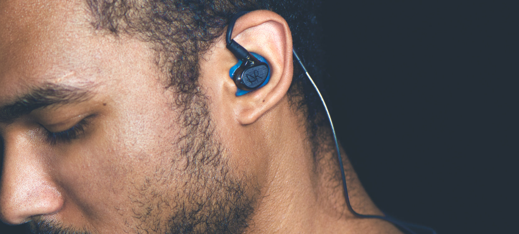 Close-up of a young man with an in-ear wireless headphone, focusing on his ear and the lower half of his face, set against a dark background.