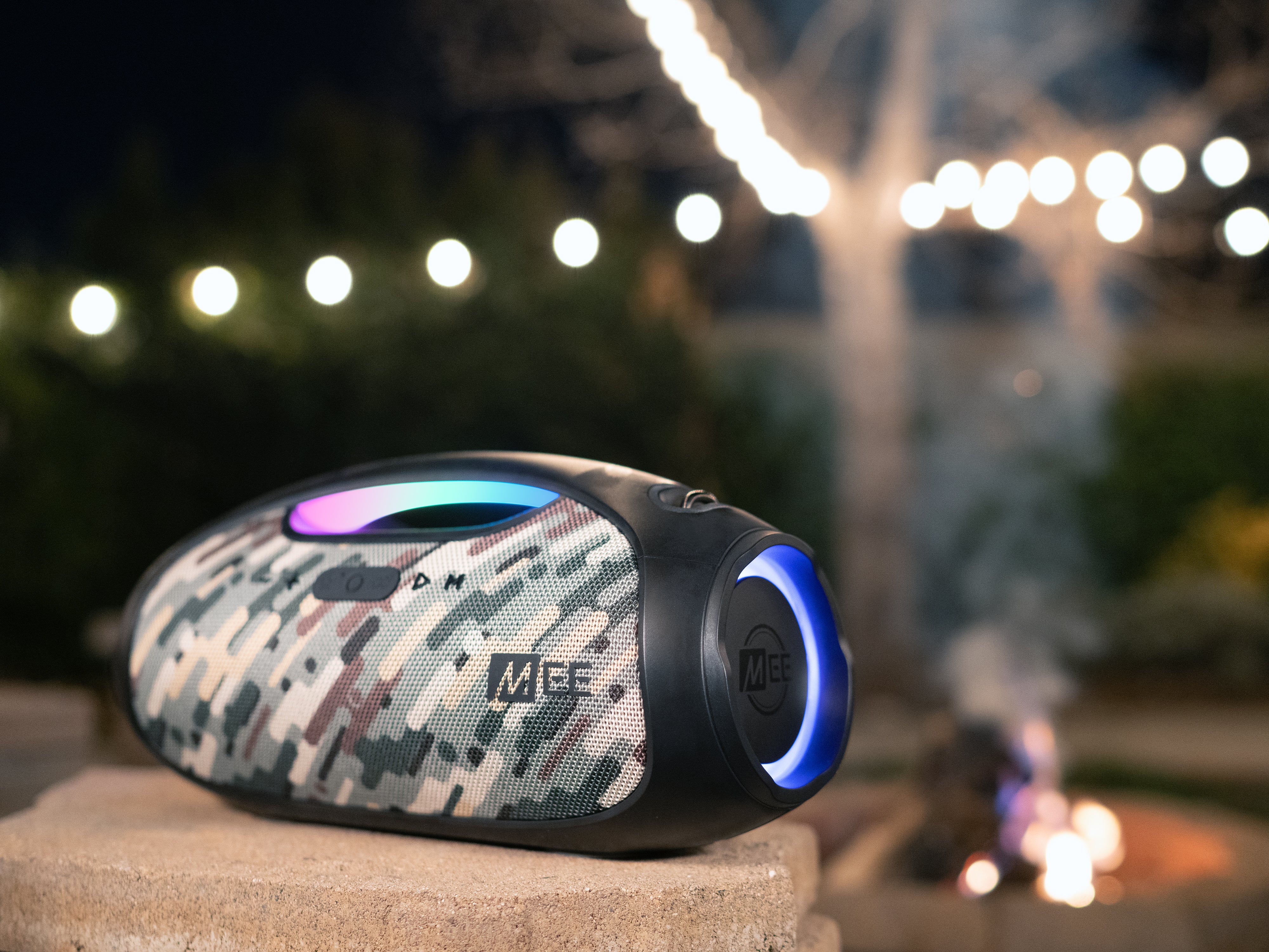 A portable speaker with a camouflage design sits on a concrete ledge, illuminated by ambient string lights and a soft glow from its circular led display, against a blurred nighttime garden backdrop.