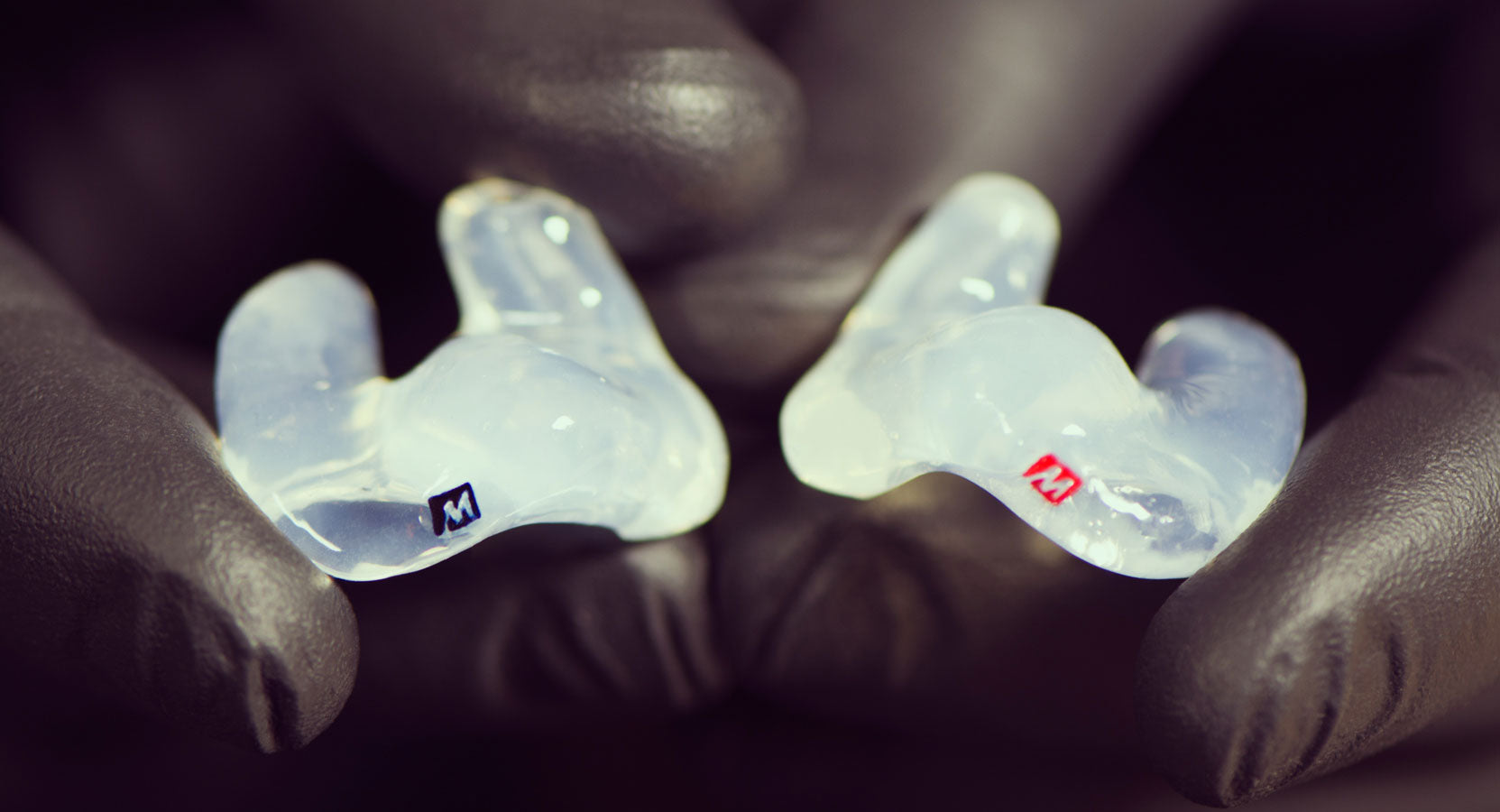 Custom earplugs held between fingers covered by a black glove. each earplug marked with an "m" on their blue and red handles respectively, suggesting left and right orientation.