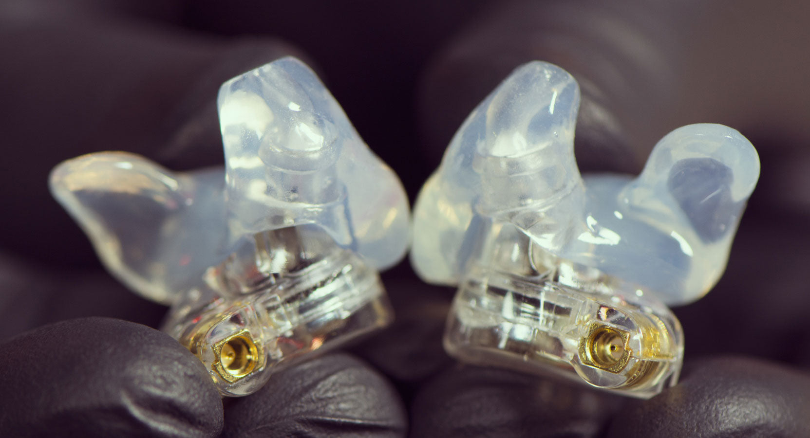 A close-up photo of a pair of custom-molded, clear earpieces held in black-gloved hands, showcasing their intricate design and transparent material.